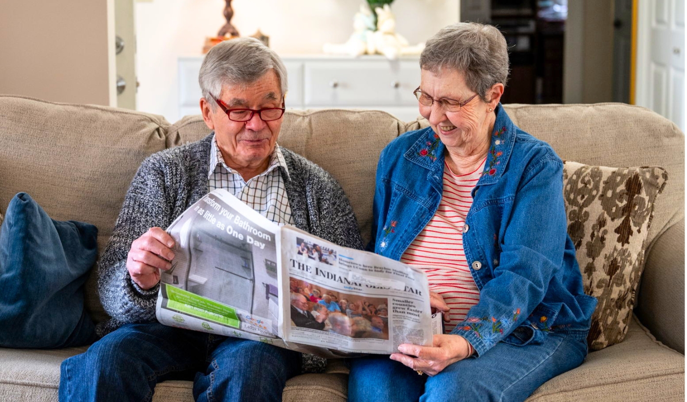 Two elderly adults sit on a beige sofa, reading a newspaper together and smiling at each other.