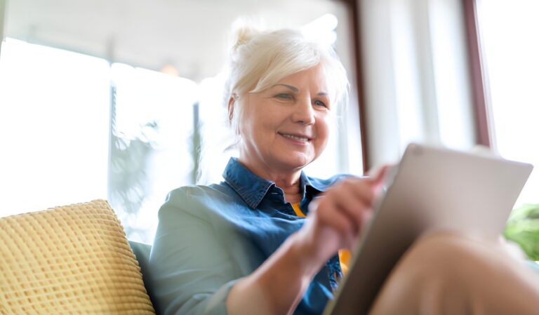 Smiling older woman on a sofa using a tablet in a bright living room.