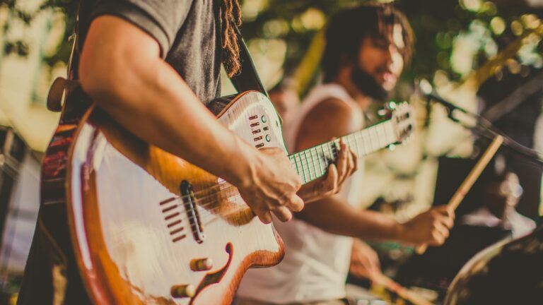 Close-up of a guitarist's hands strumming a sunburst electric guitar at an outdoor live performance, with other band members blurred in the background.