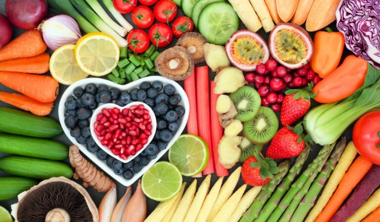 Colorful flat lay of fresh produce—fruits and vegetables arranged in a vibrant mosaic, with blueberries and pomegranate seeds in a heart-shaped bowl.