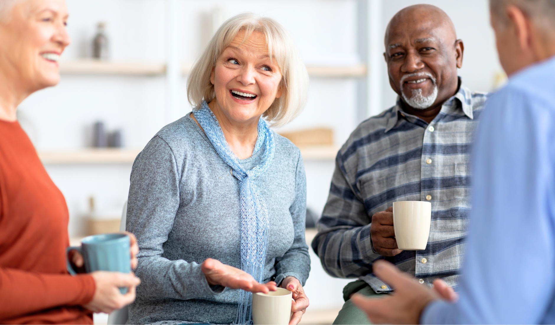 Group of older adults smiling and chatting, each holding a mug indoors in a bright room.